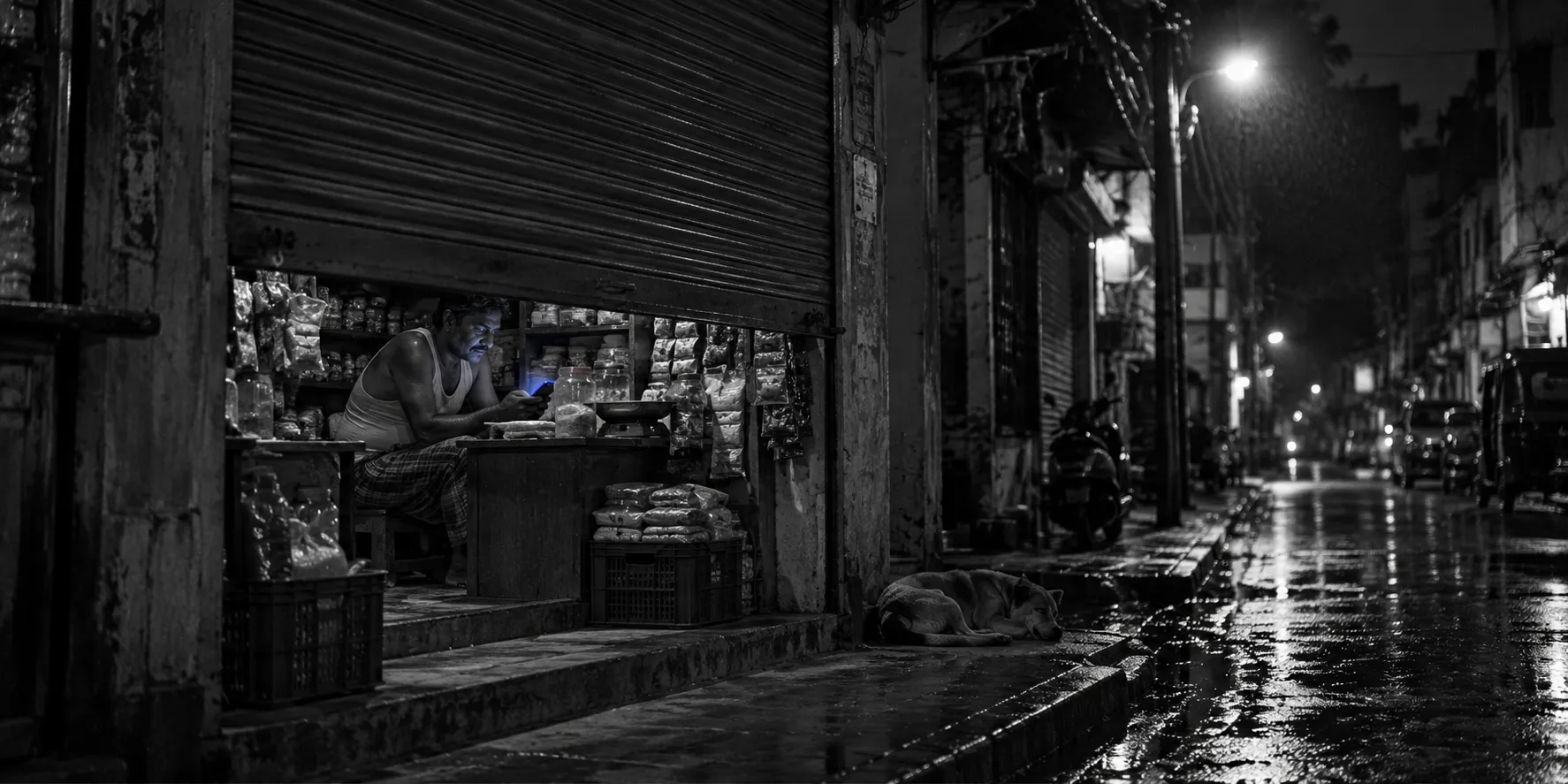 Night street with a shop shutter half down and a shopkeeper lit only by his phone
